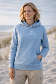 Lichtblauwe hoodie met witte lijntekening van Ameland‑vuurtorens, gedragen op strand met rustige zee