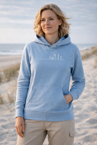 Lichtblauwe hoodie met witte lijntekening van Ameland‑vuurtorens, gedragen op strand met rustige zee