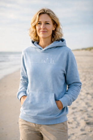 Anne op strand in lichtblauwe hoodie met witte lijntekening van Ameland‑vuurtorens, duinen en zee op achtergrond